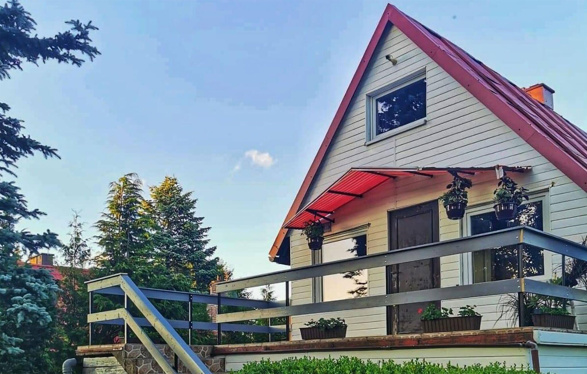 A two-story house with a red roof, a balcony with potted plants, and a stone staircase leading to the entrance.