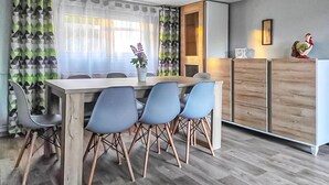 Een eetgedeelte met een houten tafel en blauwe stoelen, een potplant en een houten kastje met een decoratieve haan.