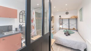 A compact hotel room with a kitchenette, a shower, a bed with a green pillow, and a small table.