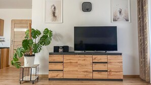 A living room with a flat-screen TV on a wooden cabinet, a potted plant, and two framed artworks on the wall.