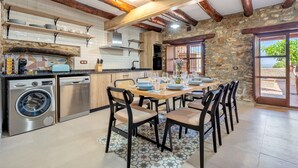 Una cocina con vigas de madera, lavadora y zona de comedor con sillas y una mesa preparada para cuatro.