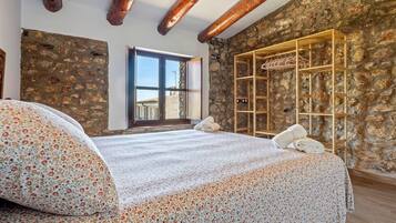 Un dormitorio con pared de piedra, vigas de madera, cama con ropa de cama floral, una ventana y un armario de madera.