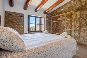 Un dormitorio con pared de piedra, vigas de madera, cama con ropa de cama floral, una ventana y un armario de madera.