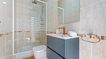 Et moderne badeværelse med brusekabine i glas, et hvidt toilet, en vask med spejl og et håndklædestativ.