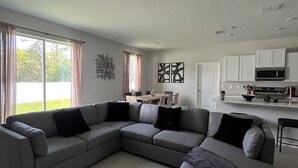 A modern living room with a sectional sofa, a dining area with a table and chairs, and a kitchen with white cabinets and appliances.