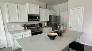 A modern kitchen with white cabinets, stainless steel appliances, a large island with stools, and a tiled floor.