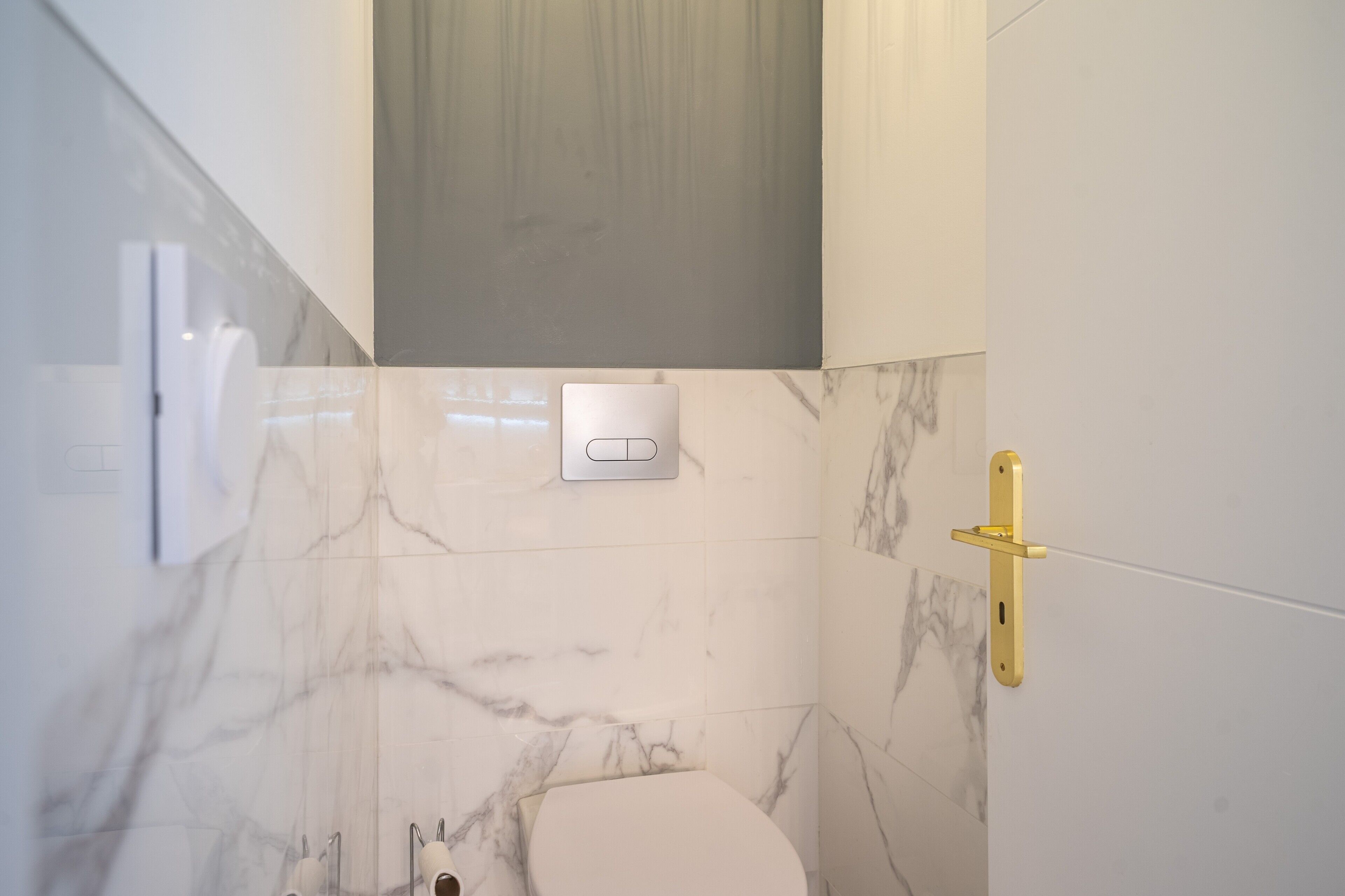 A bathroom with a toilet, a wall-mounted flush handle, and a gold-colored towel rack.