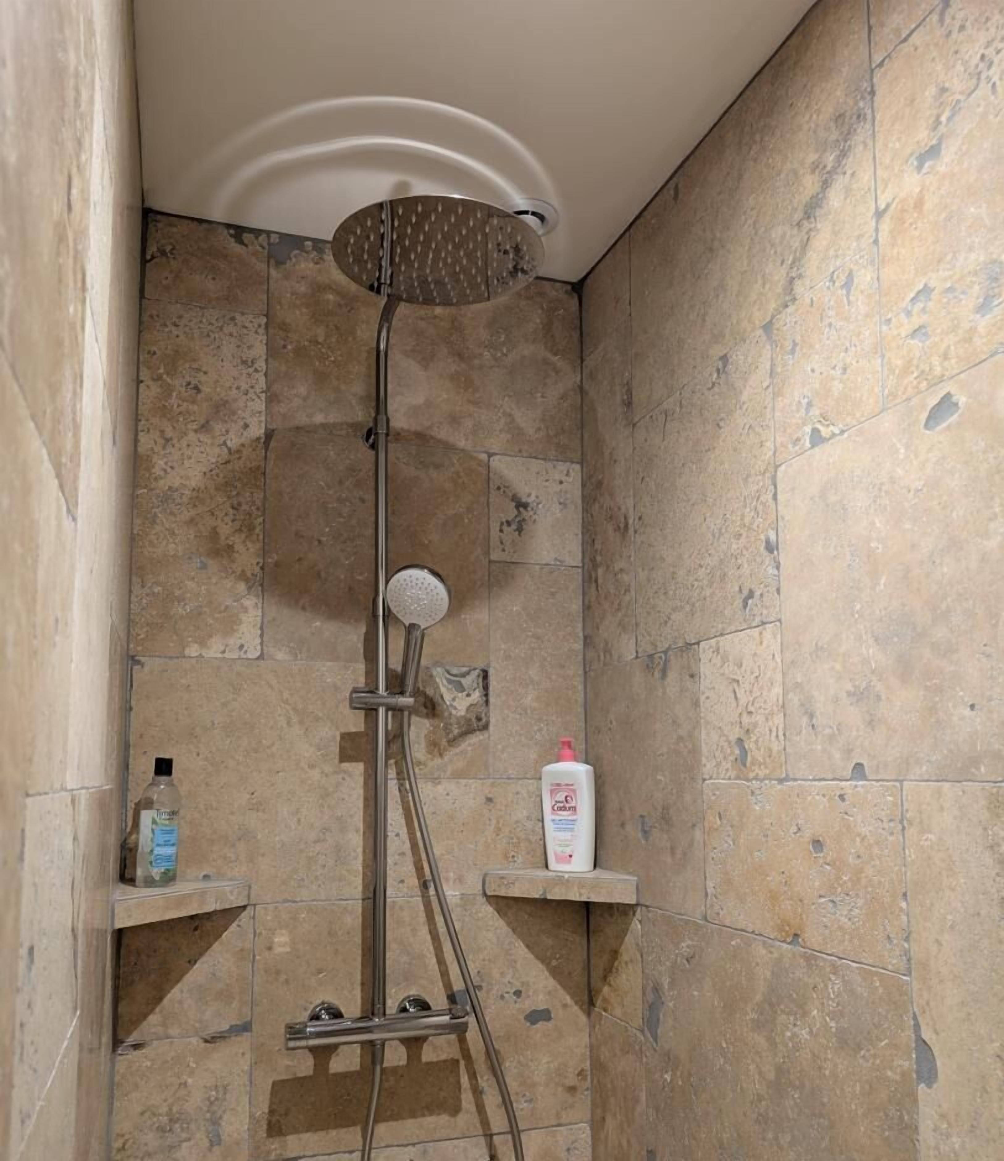 Ducha con cabezal de ducha de lluvia, cabezal de ducha manual y cepillo exfoliante. Hay dos botellas de jabón líquido en estantes montados en la pared.