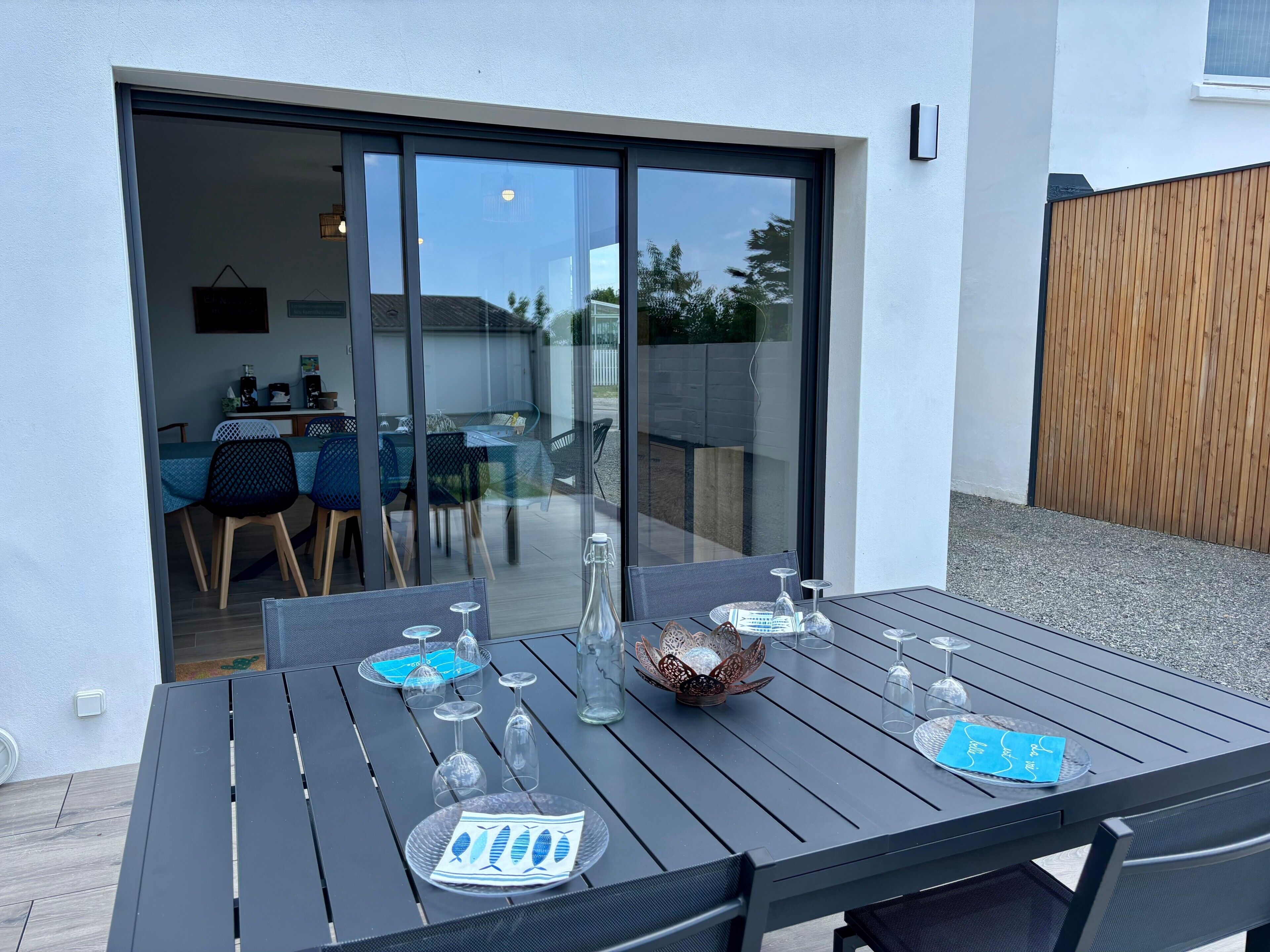 Une table de patio avec des chaises, une porte vitrée donnant sur une salle à manger intérieure et une clôture en bois.