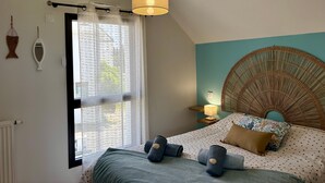 Une chambre à coucher comprenant un lit, un luminaire suspendu, une fenêtre avec des rideaux, un miroir fixé au mur et une tête de lit en osier.