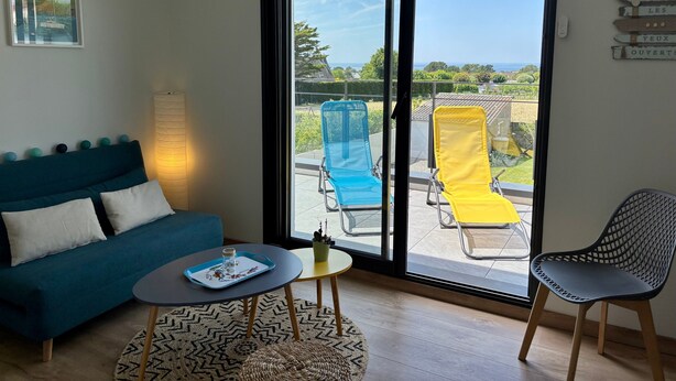 Une pièce avec un canapé bleu, une petite table ronde sur laquelle se trouve un plateau, une chaise et une vue sur un balcon équipé de deux chaises longues.