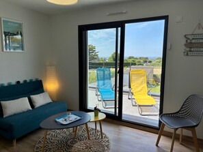 Une pièce avec un canapé bleu, une petite table ronde sur laquelle se trouve un plateau, une chaise et une vue sur un balcon équipé de deux chaises longues.
