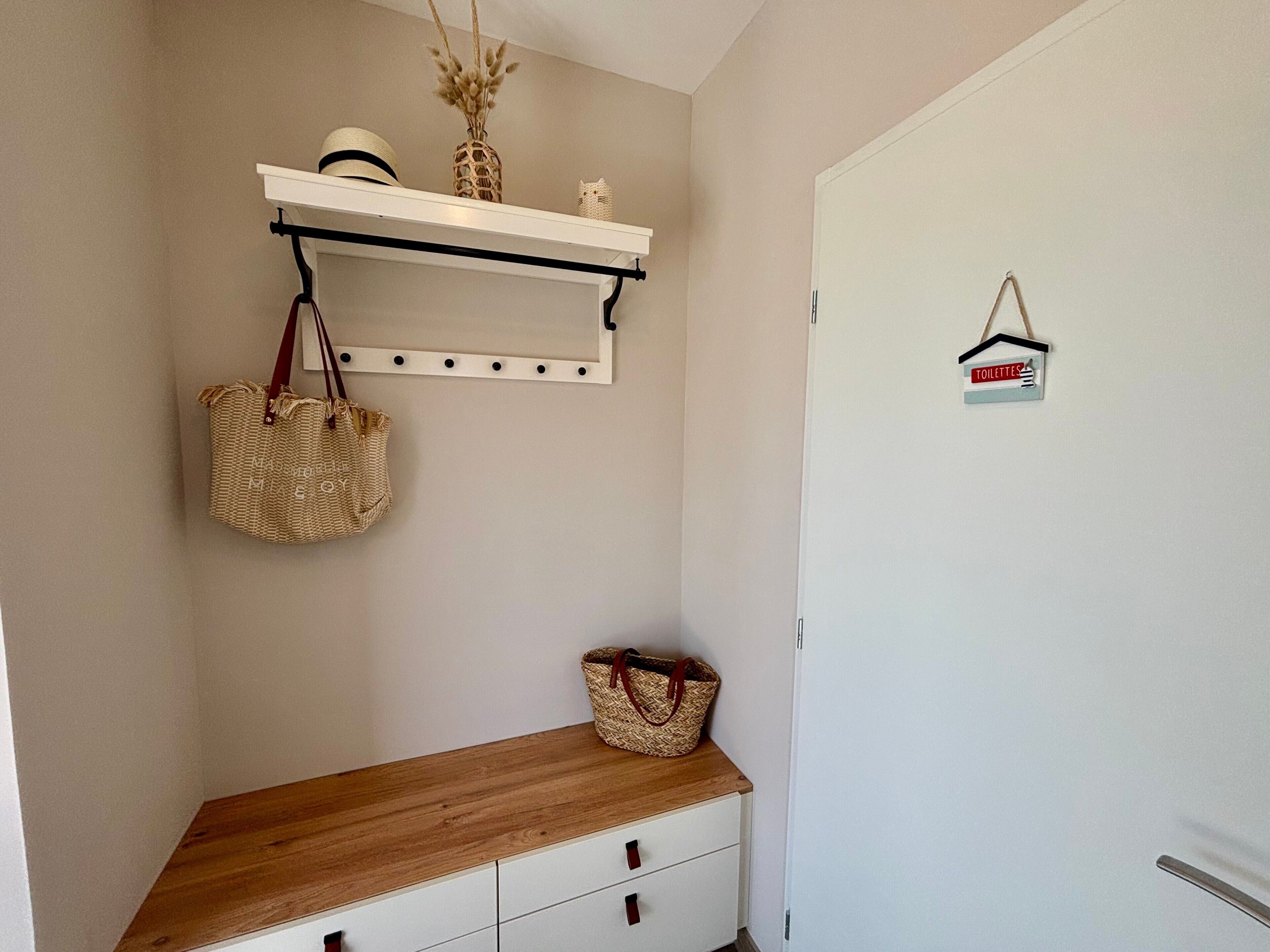 Ein Korridor mit einer Holzbank, einer hängenden Pflanzkorb, einem Wandregal mit Dekoration und einer Tür mit Hinweisschild zu den Toiletten.