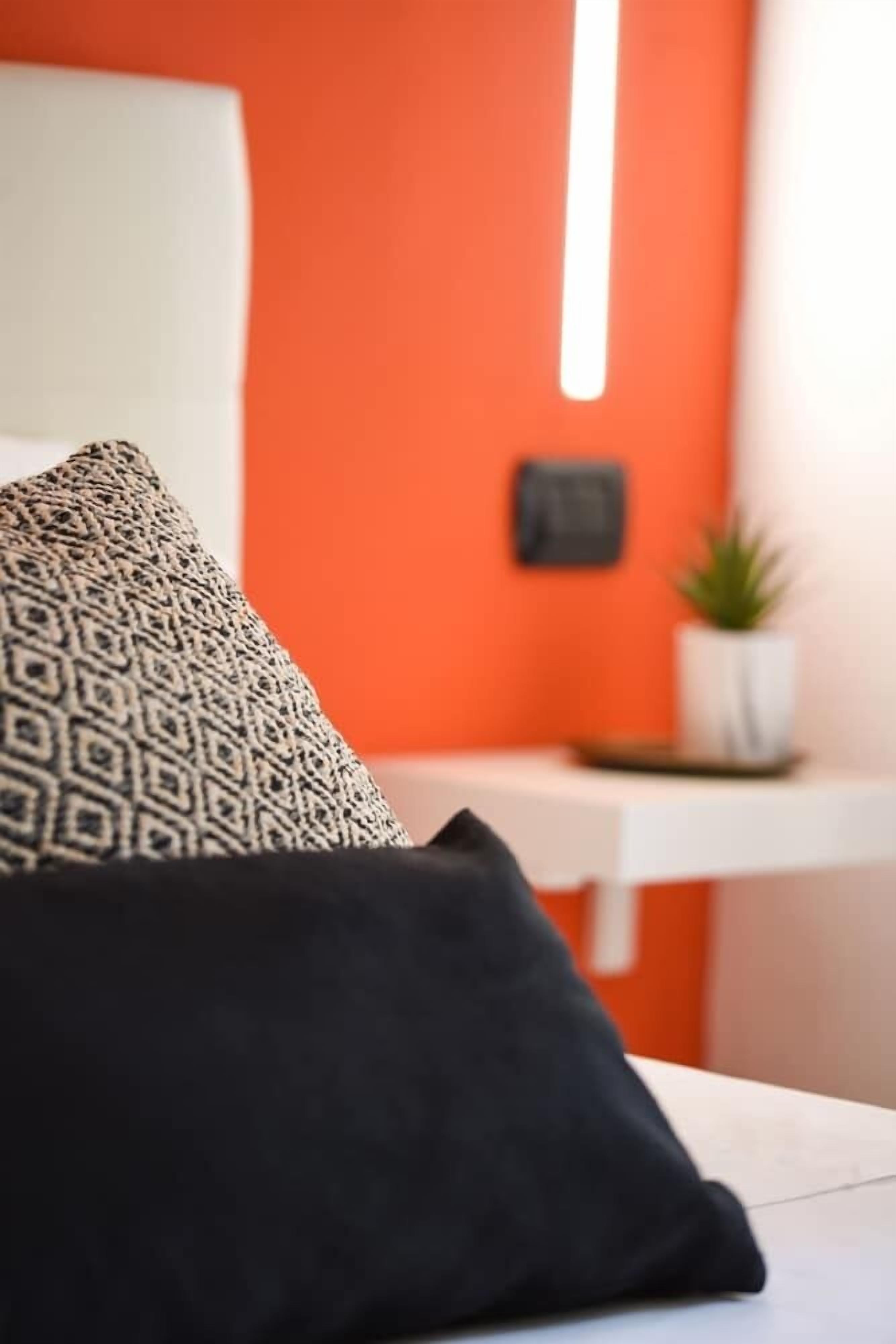 A bed with patterned and solid pillows, a white headboard, and a bright orange wall with a small plant and a framed item.