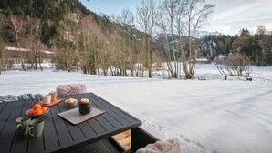 Una mesa de madera con tazas, un cuenco de naranjas y una bandeja con galletas, en un espacio exterior nevado con vistas a árboles y montañas.