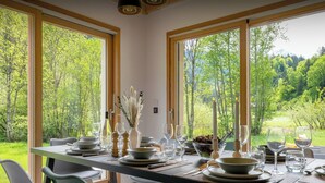 Una mesa de comedor dispuesta con platos, vasos y velas, con vistas a un paisaje boscoso a través de amplios ventanales.