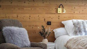 Un dormitorio acogedor con una pared de madera, una cama con ropa de cama blanca, un sillón gris mullido y una mesita de noche con un jarrón de plantas secas.