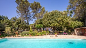 A pool with clear water, surrounded by trees and potted plants, with a building in the background.