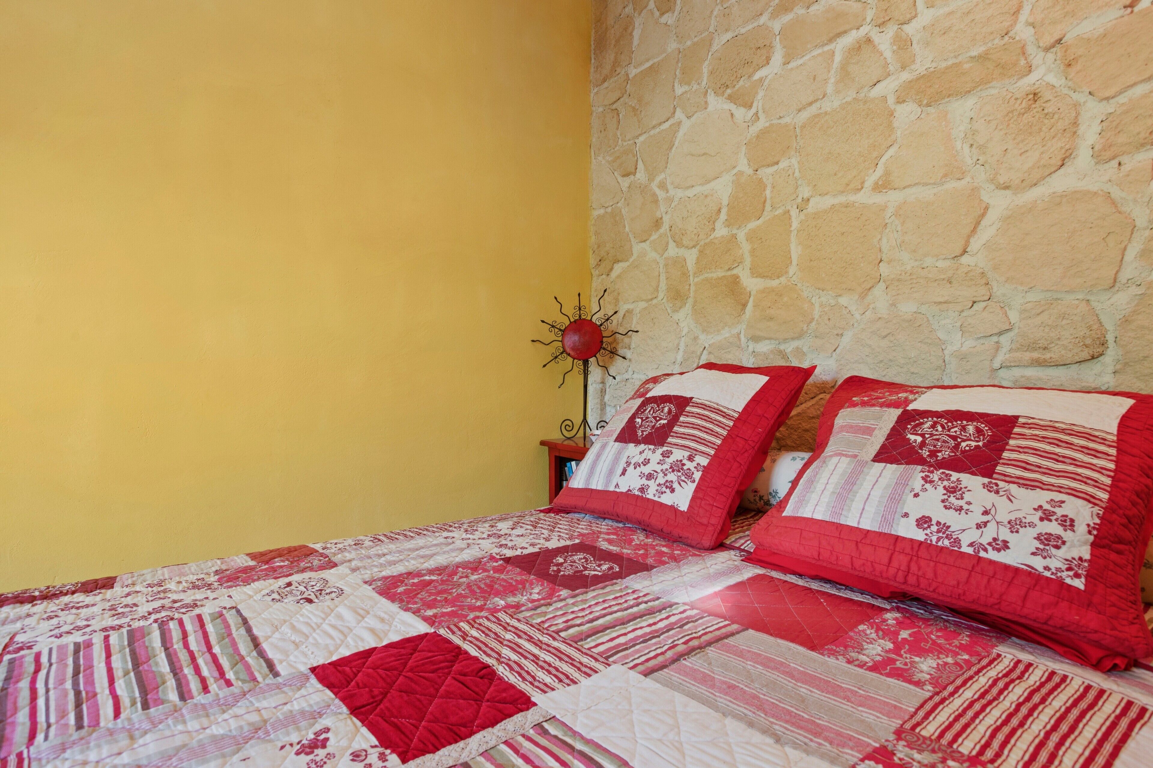 A bed with a red and white quilt, a stone wall, and a yellow wall.