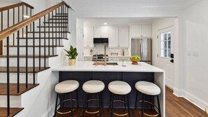 A modern kitchen with white cabinets, a central island with bar stools, a stainless steel refrigerator, and a wooden staircase.