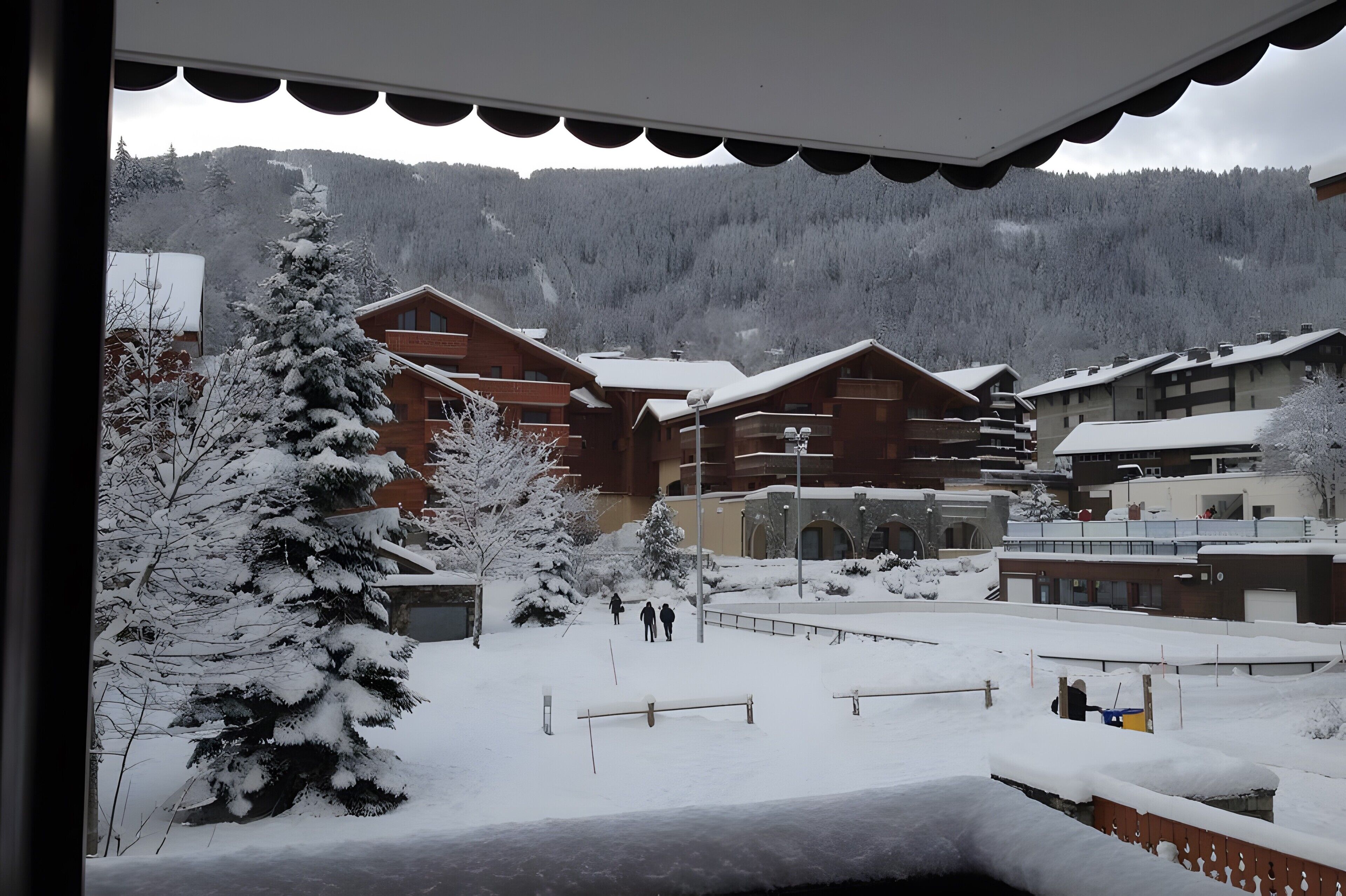 A snow-covered outdoor area with wooden buildings, a snow-covered tree, and a mountain in the background.