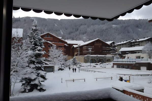 A snow-covered outdoor area with wooden buildings, a snow-covered tree, and a mountain in the background.