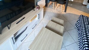 Een moderne woonkamer met een flatscreen televisie, een salontafel en een bank. Er is een eethoek met een tafel en stoelen. De keuken is zichtbaar met kasten en apparatuur.