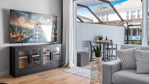 Un saló modern amb un gran televisor de pantalla plana, un sofà gris, una zona de menjador amb taula i cadires, i vista d'un edifici a través d'una finestra gran.