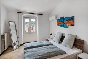 Une chambre à coucher comprenant un lit, une fenêtre, un radiateur, un miroir, une table de chevet et un climatiseur fixé au mur.