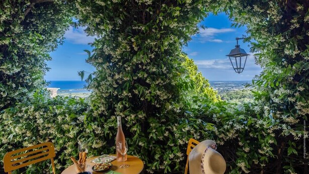 Una mesa dispuesta con una botella, vasos y aperitivos, con vistas a un paisaje pintoresco, una lámpara y abundante follaje.