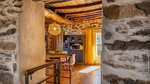 Un interior rústico con paredes de piedra vista, techo de madera, zona de comedor con mesa y sillas, y cocina con armarios y una lámpara colgante.