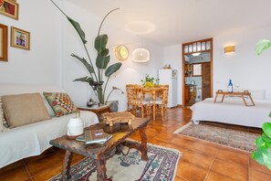 Una sala de estar con sofá, mesa de centro y mesa de comedor. Se observa una planta grande, una lámpara y un refrigerador al fondo.