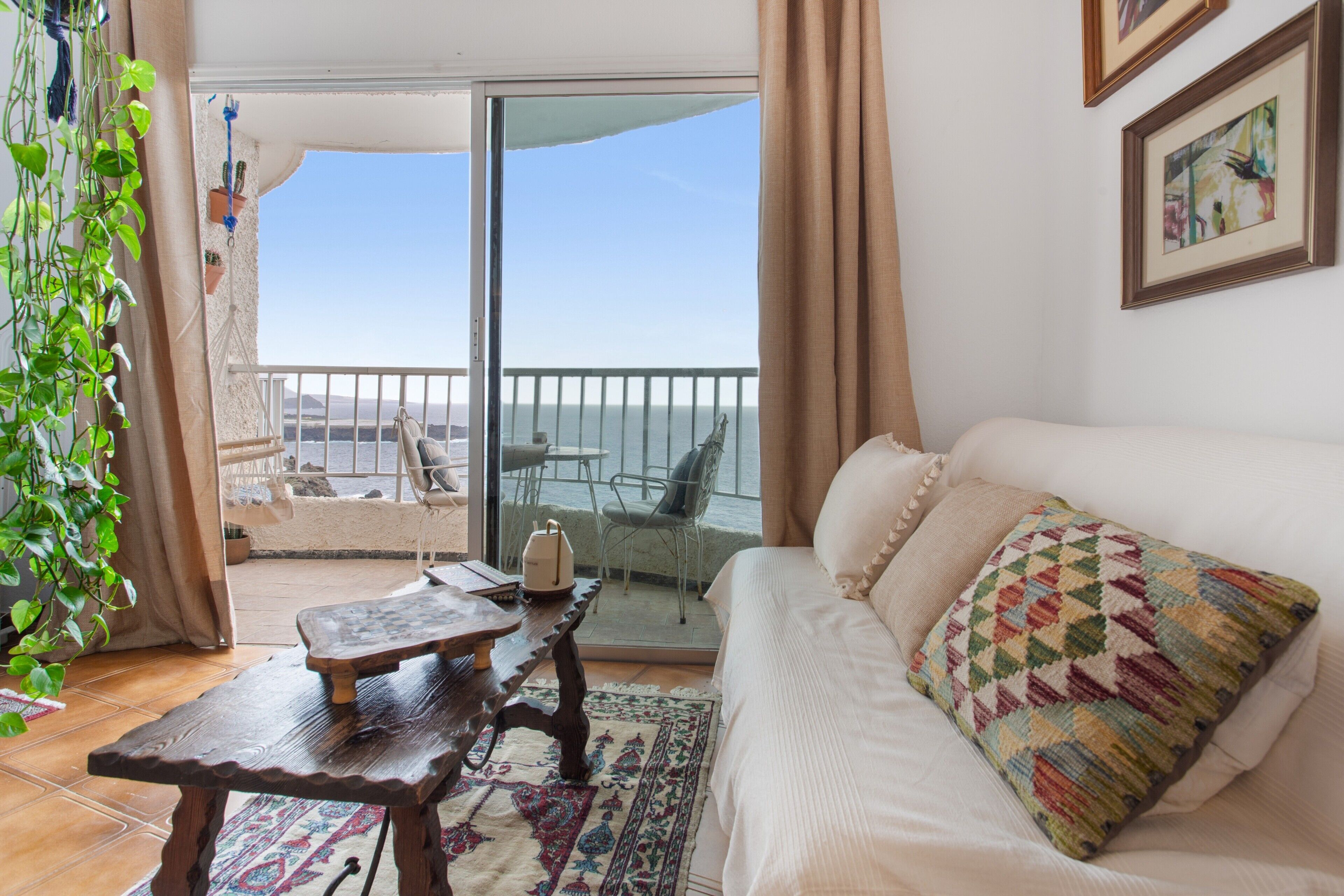 Une pièce comprenant un canapé, une table en bois, un tapis à motifs, une plante et une vue sur la mer par une porte vitrée.