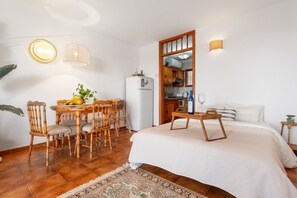 Un dormitorio con cama, mesita de noche y una pequeña mesa con una silla. Cuenta con una zona de cocina con armarios y un refrigerador.
