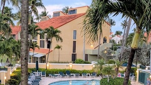 Poolområde i et resort med liggestole og parasoller, omgivet af palmer og en bygning med rødt tag.