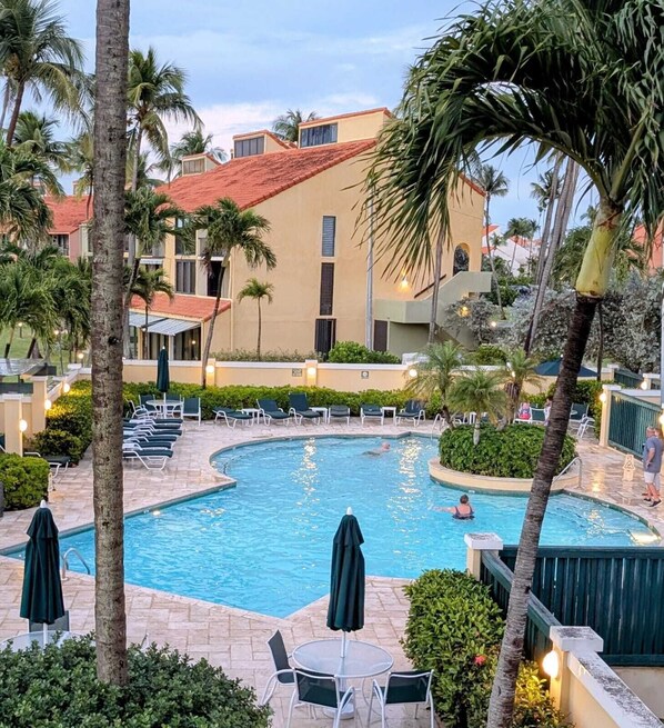 A resort pool area with lounge chairs and umbrellas, surrounded by palm trees and a building with a red roof.