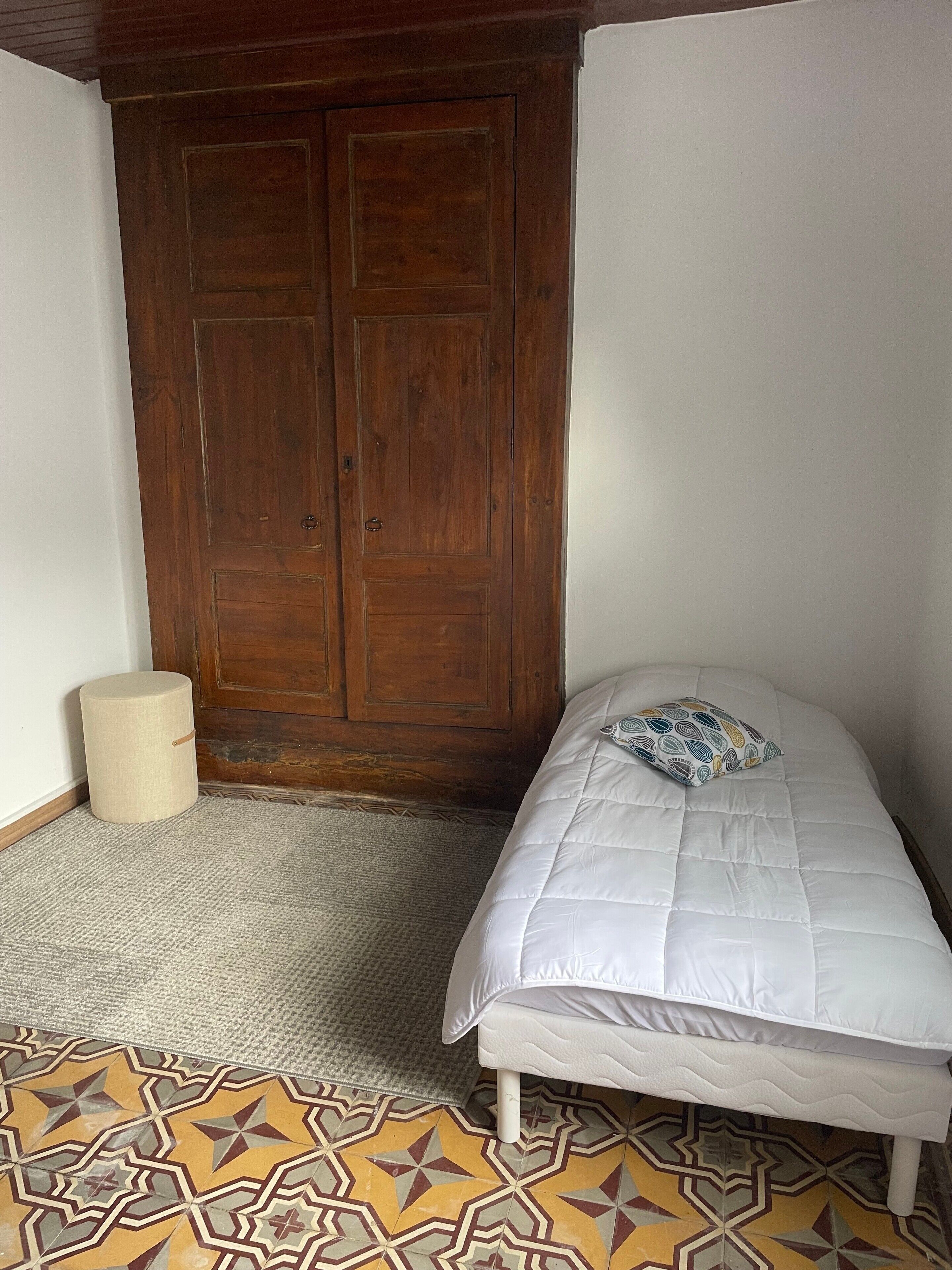 A bedroom with a bed, a wooden wardrobe, a small round stool, and patterned floor tiles.