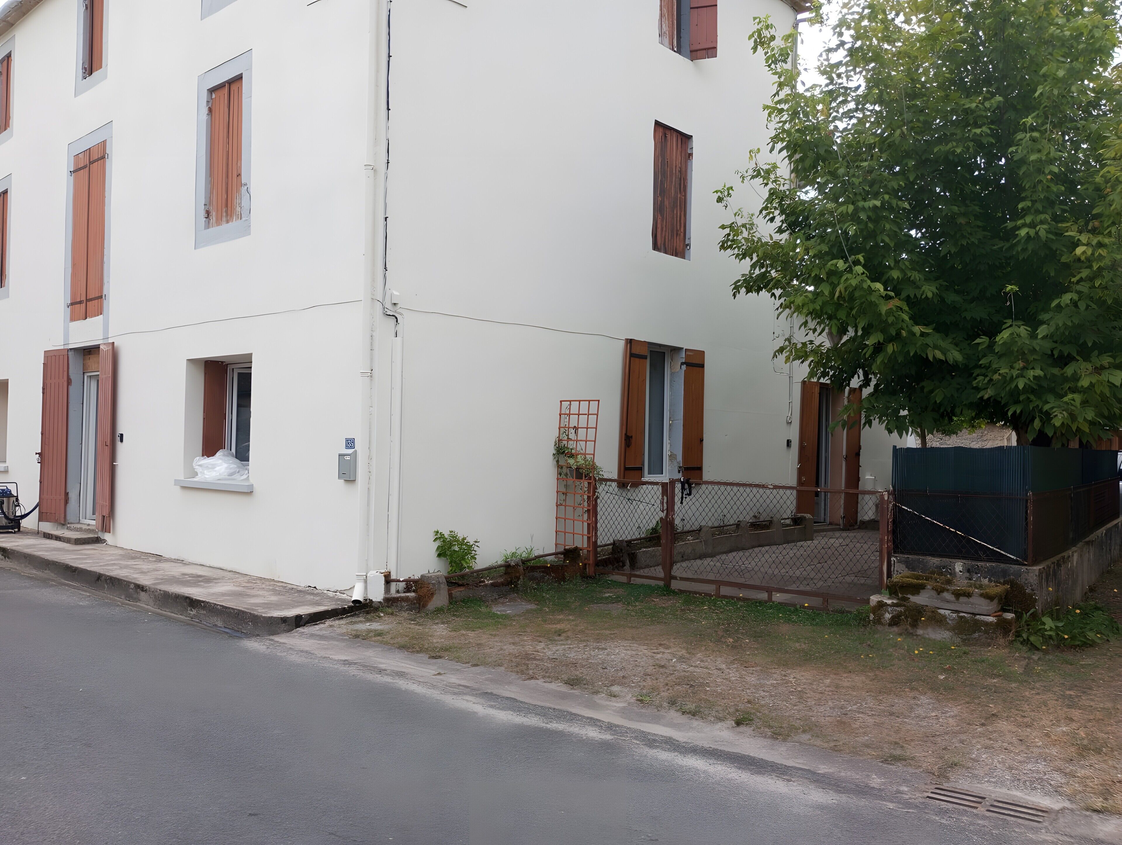 Un immeuble à plusieurs étages, doté d’une façade blanche, de nombreuses fenêtres avec des volets en bois, et d’un petit espace extérieur agrémenté d’une clôture en métal et de quelques marches.