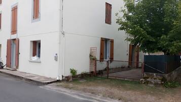 Un immeuble à plusieurs étages, doté d’une façade blanche, de nombreuses fenêtres avec des volets en bois, et d’un petit espace extérieur agrémenté d’une clôture en métal et de quelques marches.