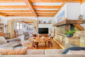 Un salon chaleureux avec une cheminée en pierre, des poutres en bois, un téléviseur et des fauteuils confortables.