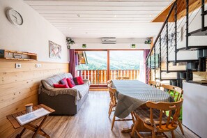 A cozy room with a wooden floor, a sofa with cushions, a dining table with chairs, a balcony with a view, and a staircase.
