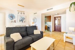 A living room with a sofa, a coffee table, and a dining area with chairs. There is a kitchen visible in the background. - Golf Relax In Betanzos Apartment with Shared Pool, Wi-Fi, and Air Conditioning (Fuente Alamo)