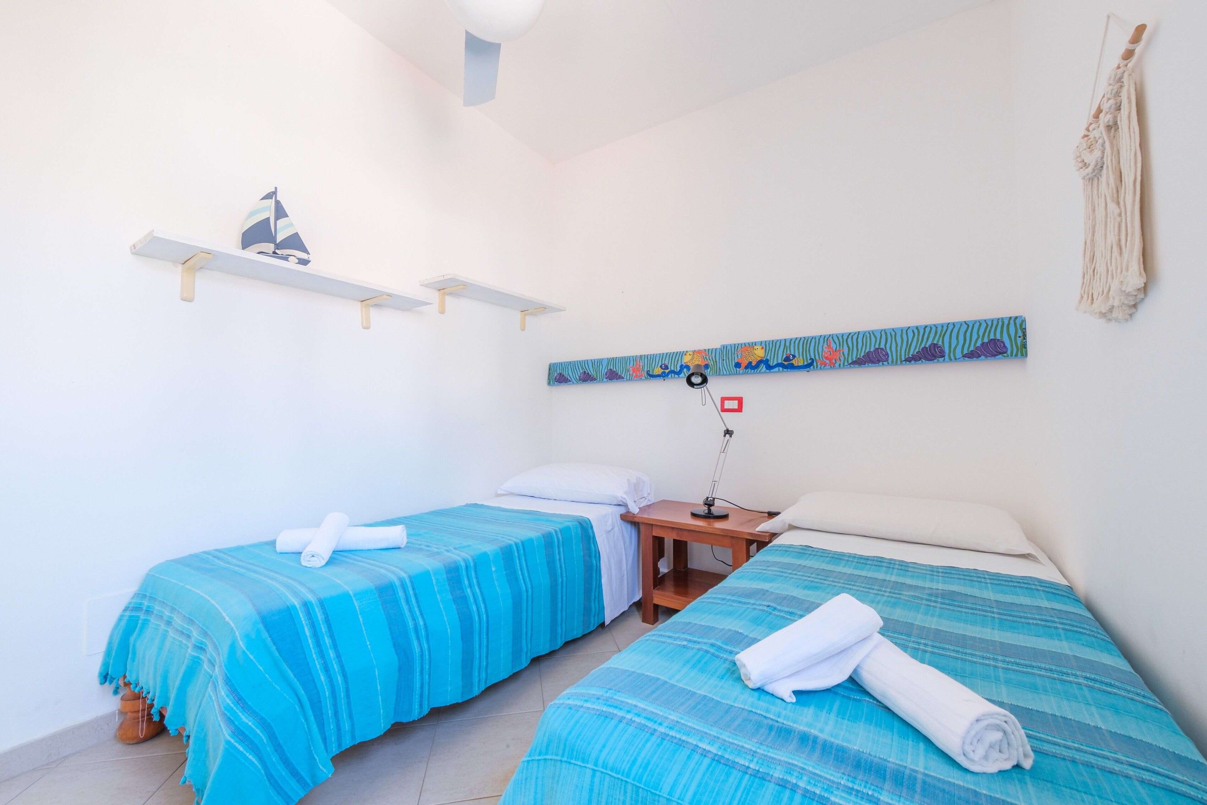 A room with two beds, a nightstand, a wall-mounted shelf with a sailboat decoration, and a macrame wall hanging.