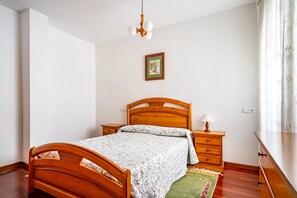 Un dormitorio con cama de madera, mesita de noche, cómoda, un cuadro enmarcado en la pared y una ventana con cortinas.