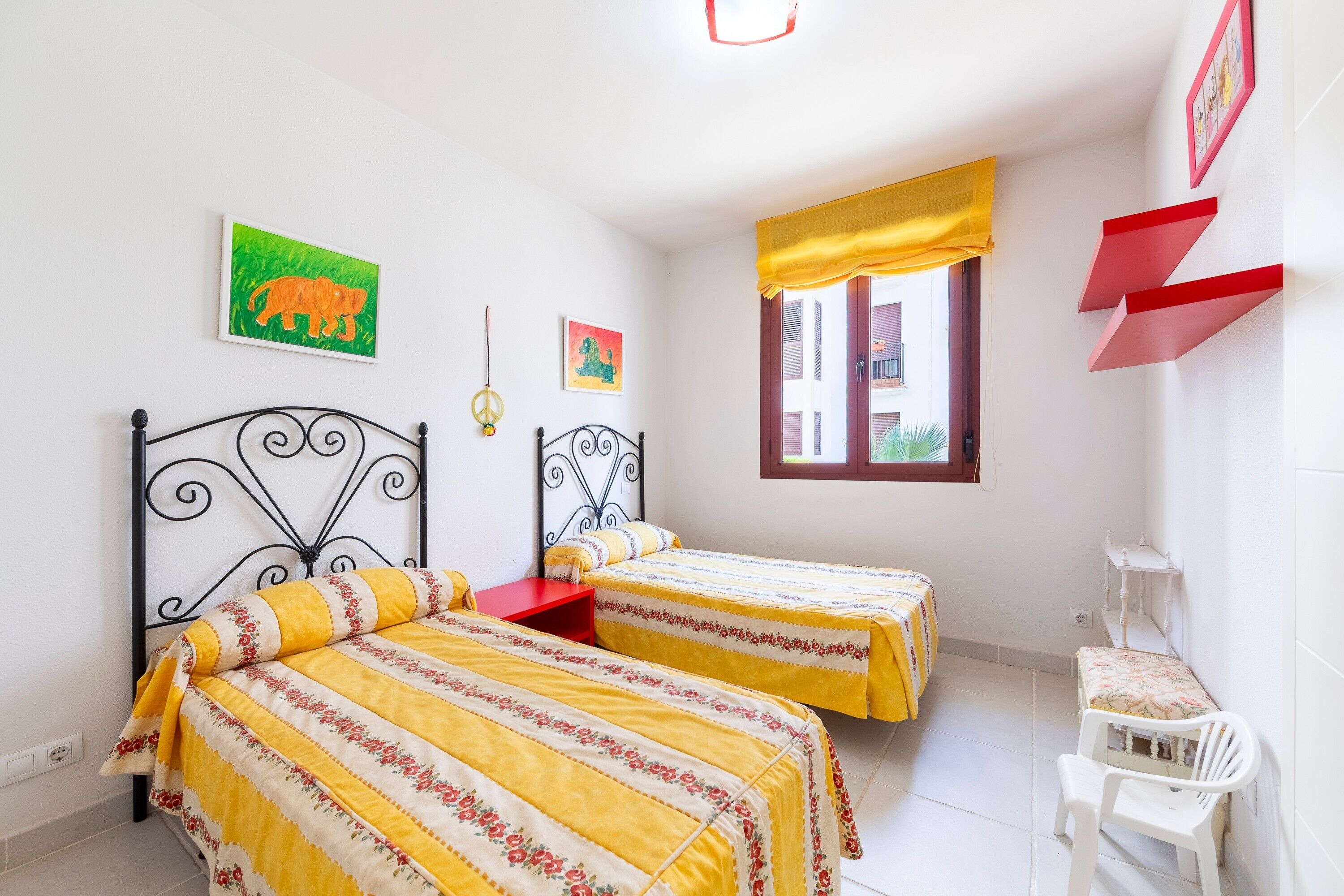 A bedroom with two beds, a window with yellow curtains, and a small white chair.