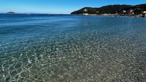 A clear beach with visible pebbles and calm water, a distant island, and a hilly landscape with buildings.