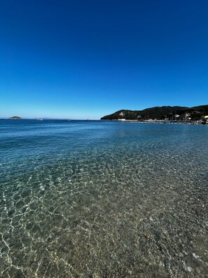 Ein unberührter Strand mit sichtbaren Kieselsteinen und ruhigem Wasser, eine entfernte Insel und eine hügelige Landschaft mit Gebäuden.