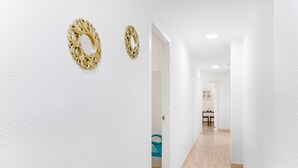 A hallway with white walls and wooden flooring, leading to a room with a bed and a table.