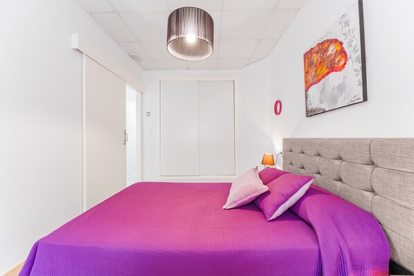 A bedroom with a bed, a headboard, a wall-mounted lamp, a door, and a painting.