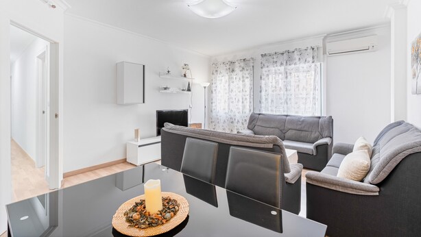 A modern living room with a glass dining table, grey sofa, and a flat-screen TV.